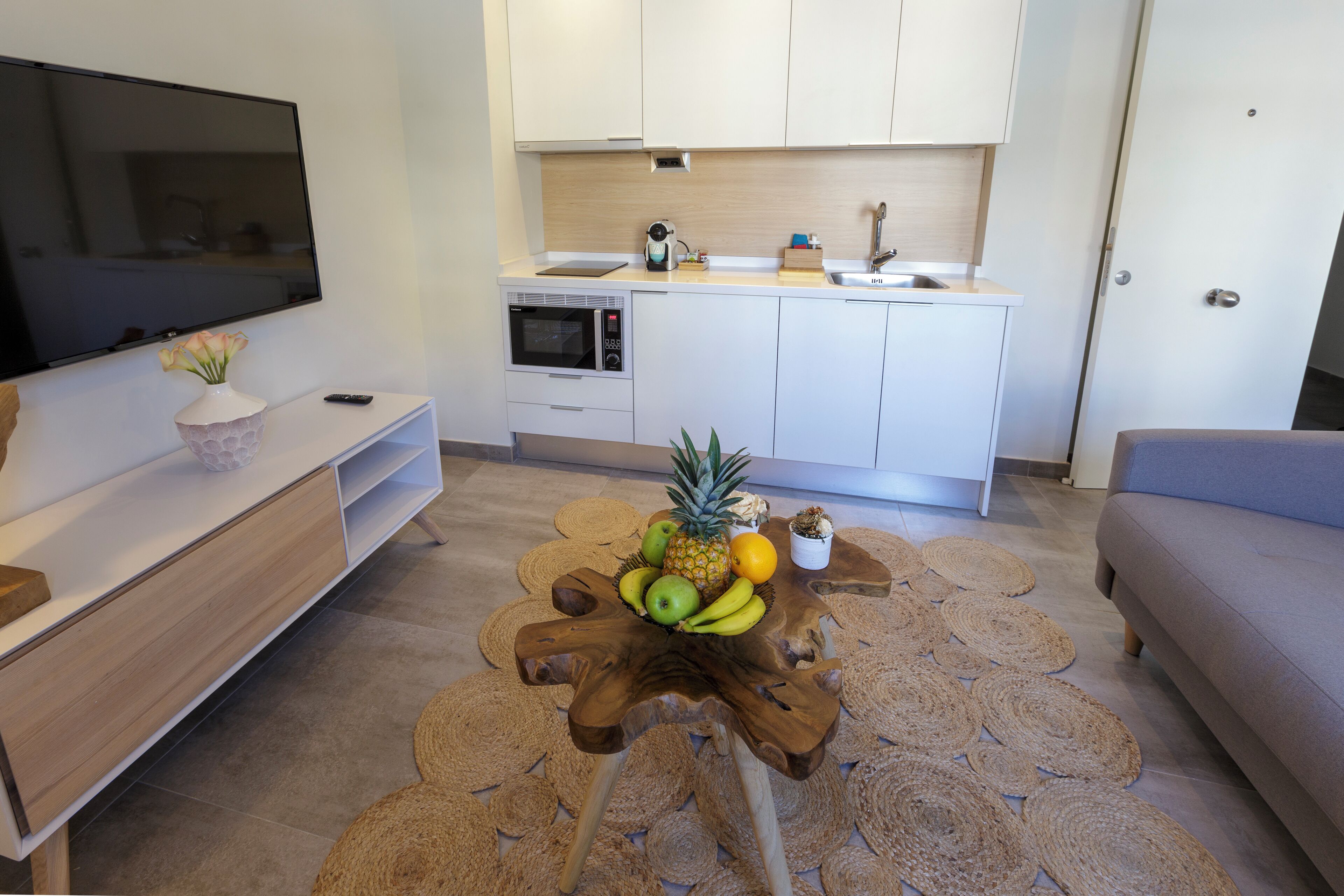 A compact living space with a kitchenette, a flat-screen TV, a wooden coffee table with fruits, and a grey sofa.