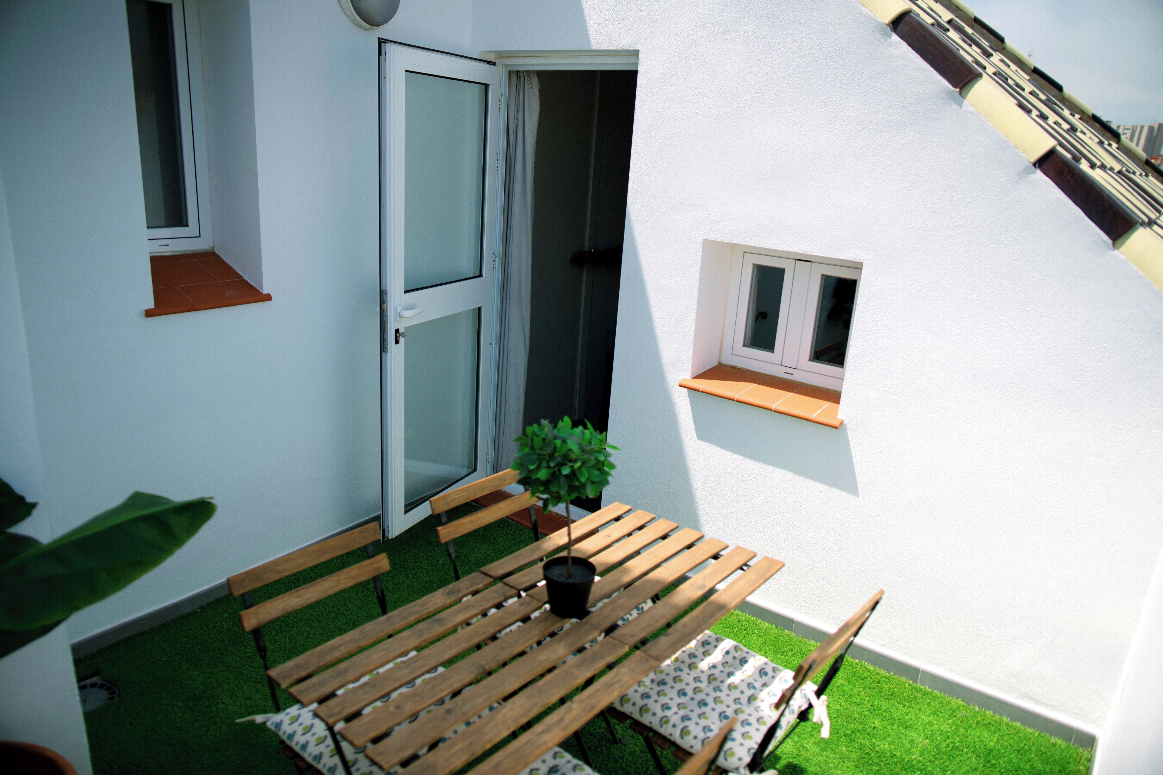 Ein kleiner Außenbereich mit Holztisch und Bänken, einem Blumentopf und einem kleinen Fenster.