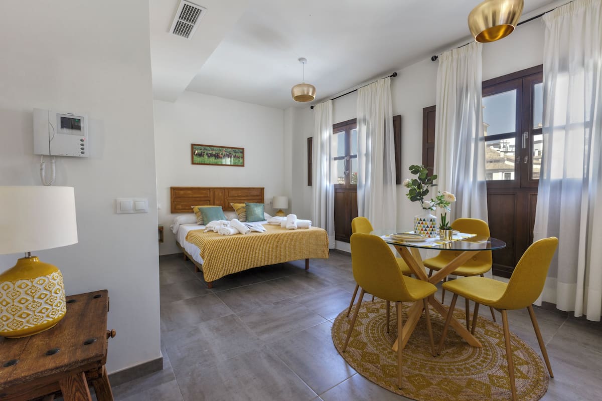 A hotel room with a bed, a bedside table with a lamp, a dining area with a table and chairs, and a window with curtains.
