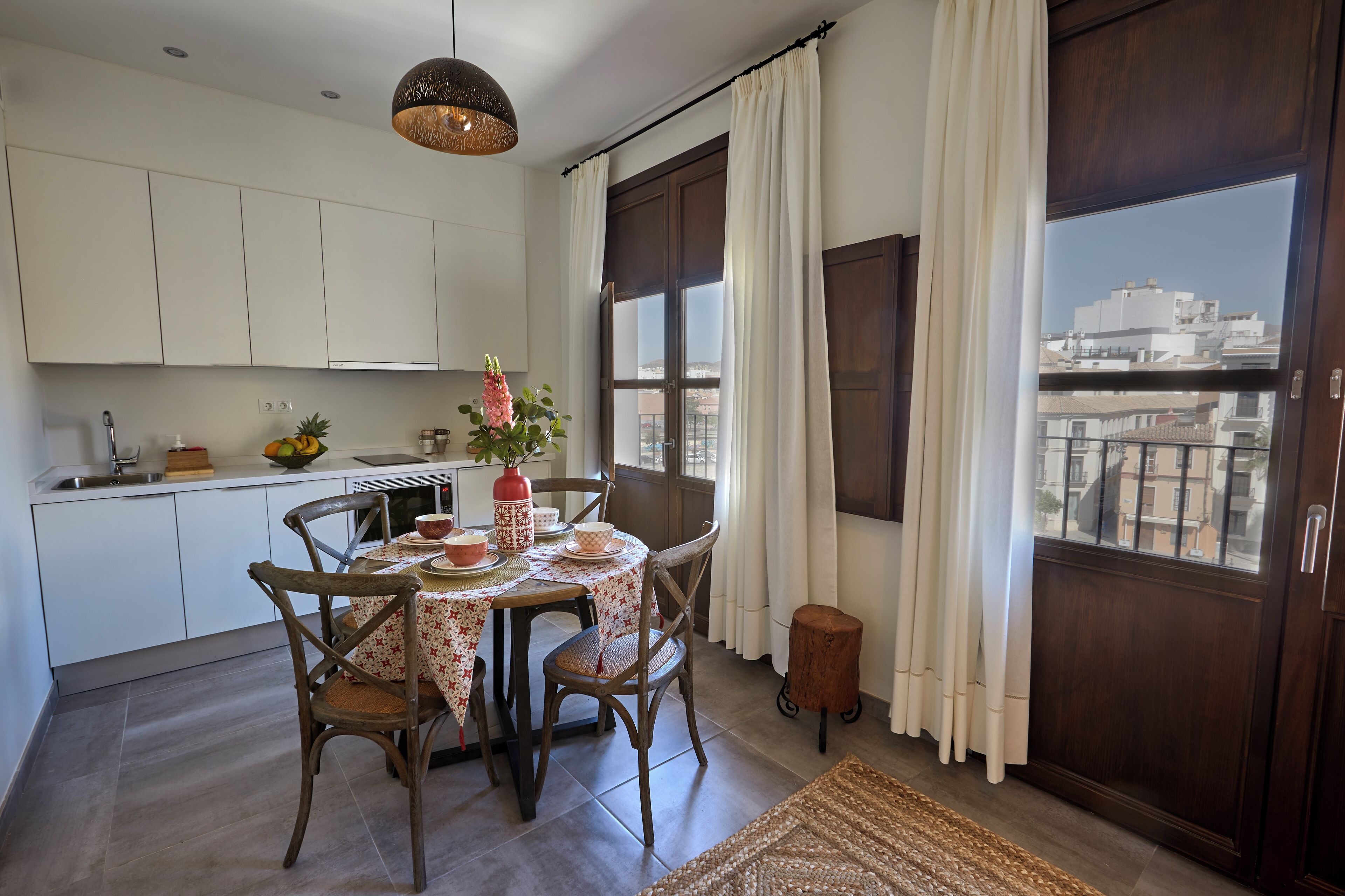 A kitchen with white cabinets, a dining table with chairs, a hanging light fixture, and a view of buildings through the window.