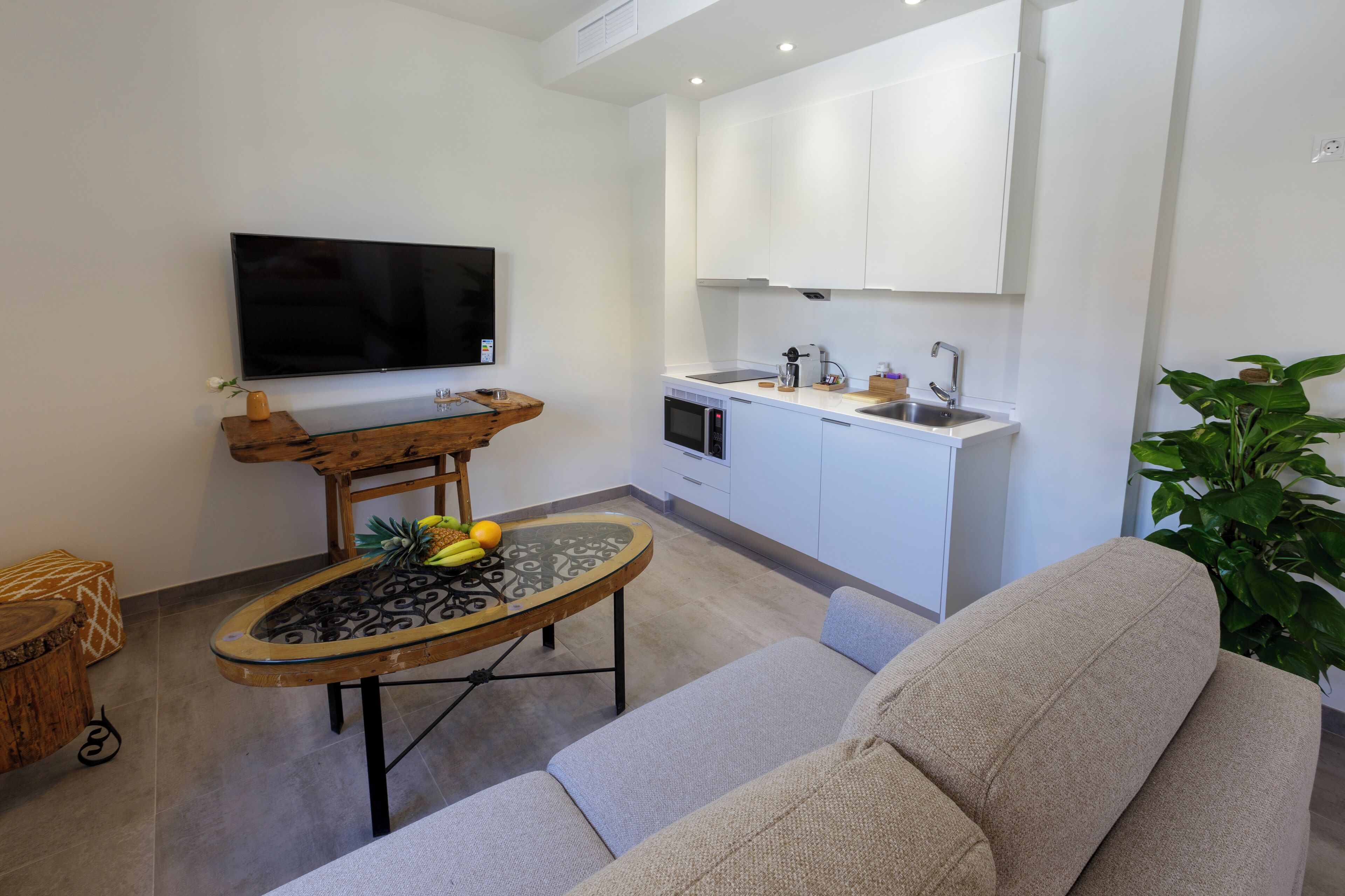 Una cocina moderna con armarios blancos, horno empotrado y microondas. Hay una mesa de comedor con cubierta de vidrio y una mesita auxiliar de madera. Un televisor de pantalla plana está montado en la pared.