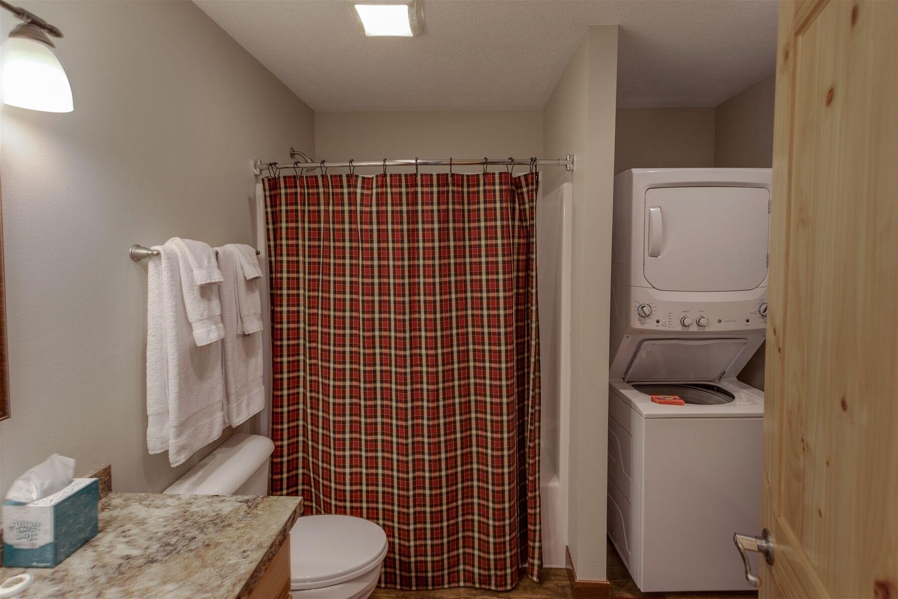 Une petite salle de bain comprenant des toilettes, un lavabo avec un plan de travail en marbre, un rideau de douche à carreaux et un lave-linge à chargement frontal.