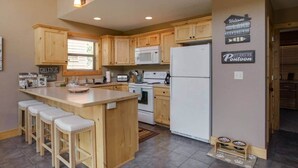 A kitchen with wooden cabinets, a central island with stools, a refrigerator, and a microwave. - Cabin 109 - Open Loft/ Modern/Clean/Comfortable (Walker)