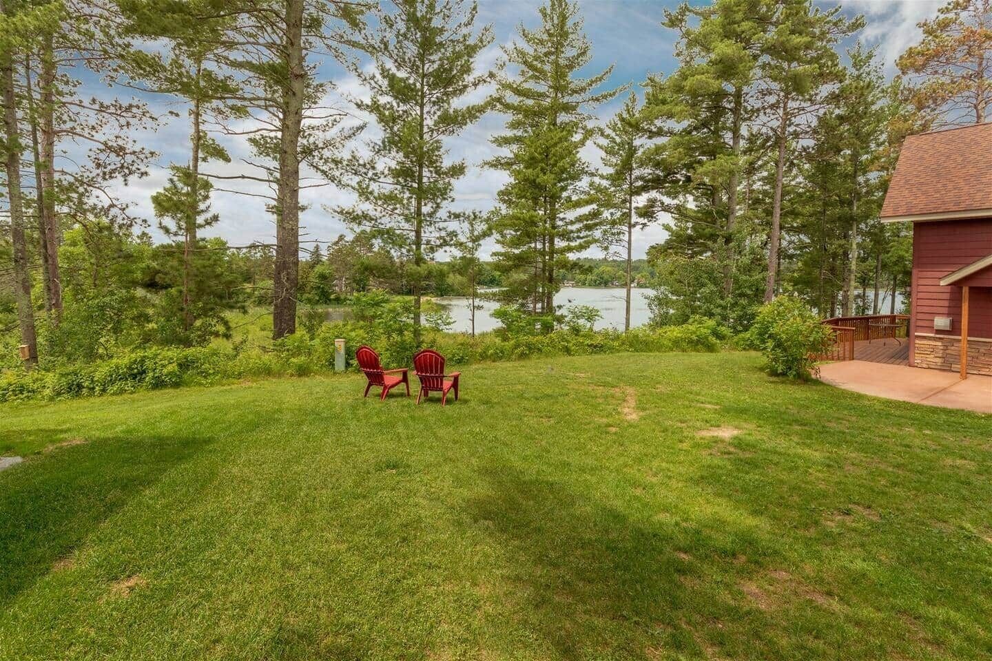 Une cour verdoyante avec deux fauteuils Adirondack, une terrasse en bois et une vue sur un lac.