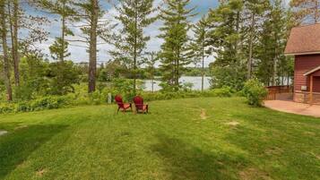 Une cour verdoyante avec deux fauteuils Adirondack, une terrasse en bois et une vue sur un lac.