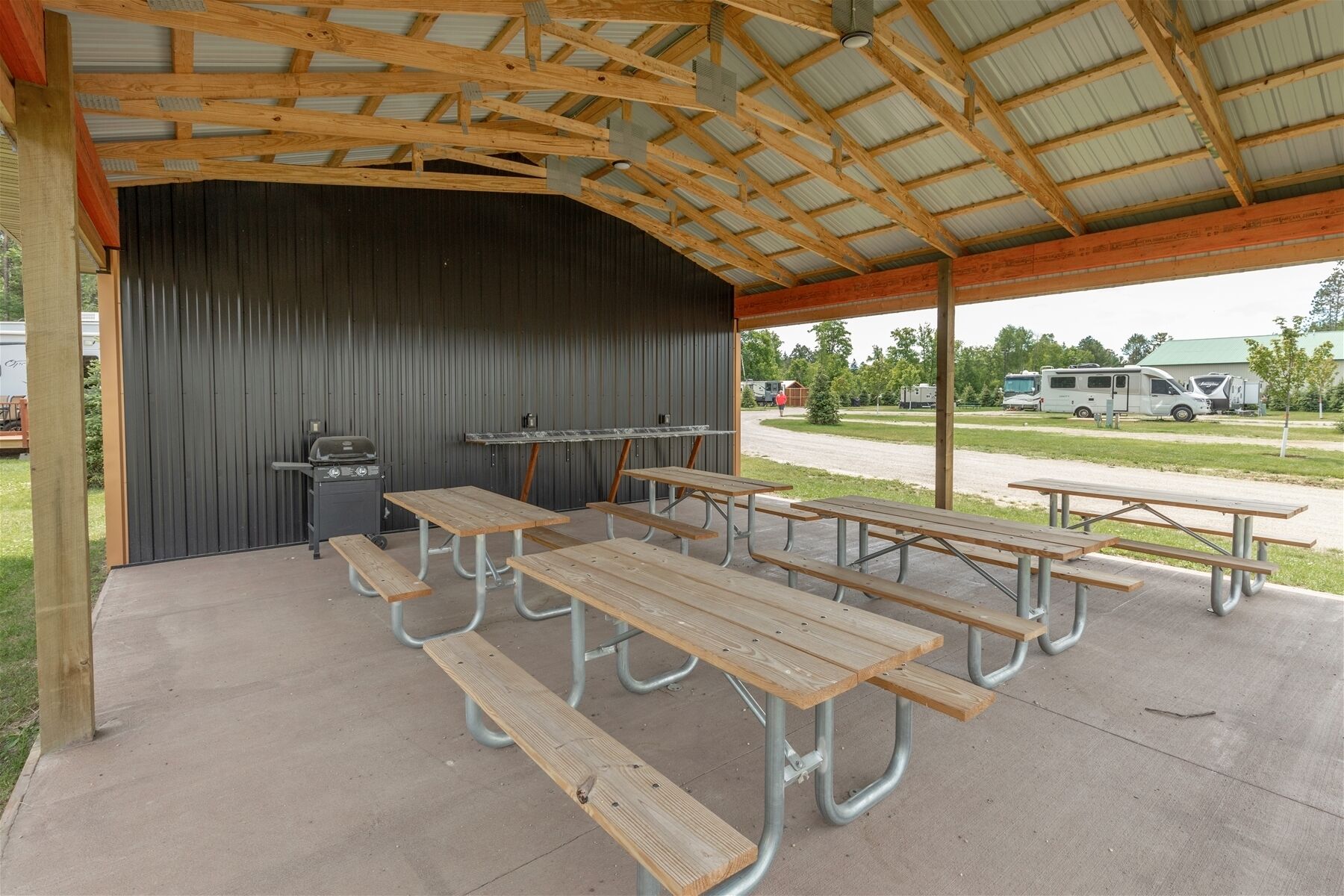 An outdoor covered picnic area with metal tables and benches, a grill, and a view of a parking lot with RVs.