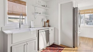 A kitchen with white cabinetry, a stainless steel sink, and a dishwasher. There is a window with a bamboo blind, and a door leading to another room. - Linden Hills Edina. Remolded Home Near the Lakes. King Bed. Sleeps 10. (Minneapolis)