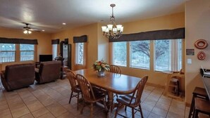 A dining area with a wooden table and chairs, a chandelier, and large windows with blinds.