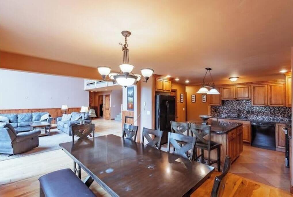 A spacious living room with a dark wooden dining table and chairs, a large sofa set, and a kitchen area with wooden cabinets and granite countertops.