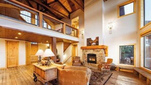 A spacious living room with a stone fireplace, wooden floors, and a high ceiling with exposed beams.