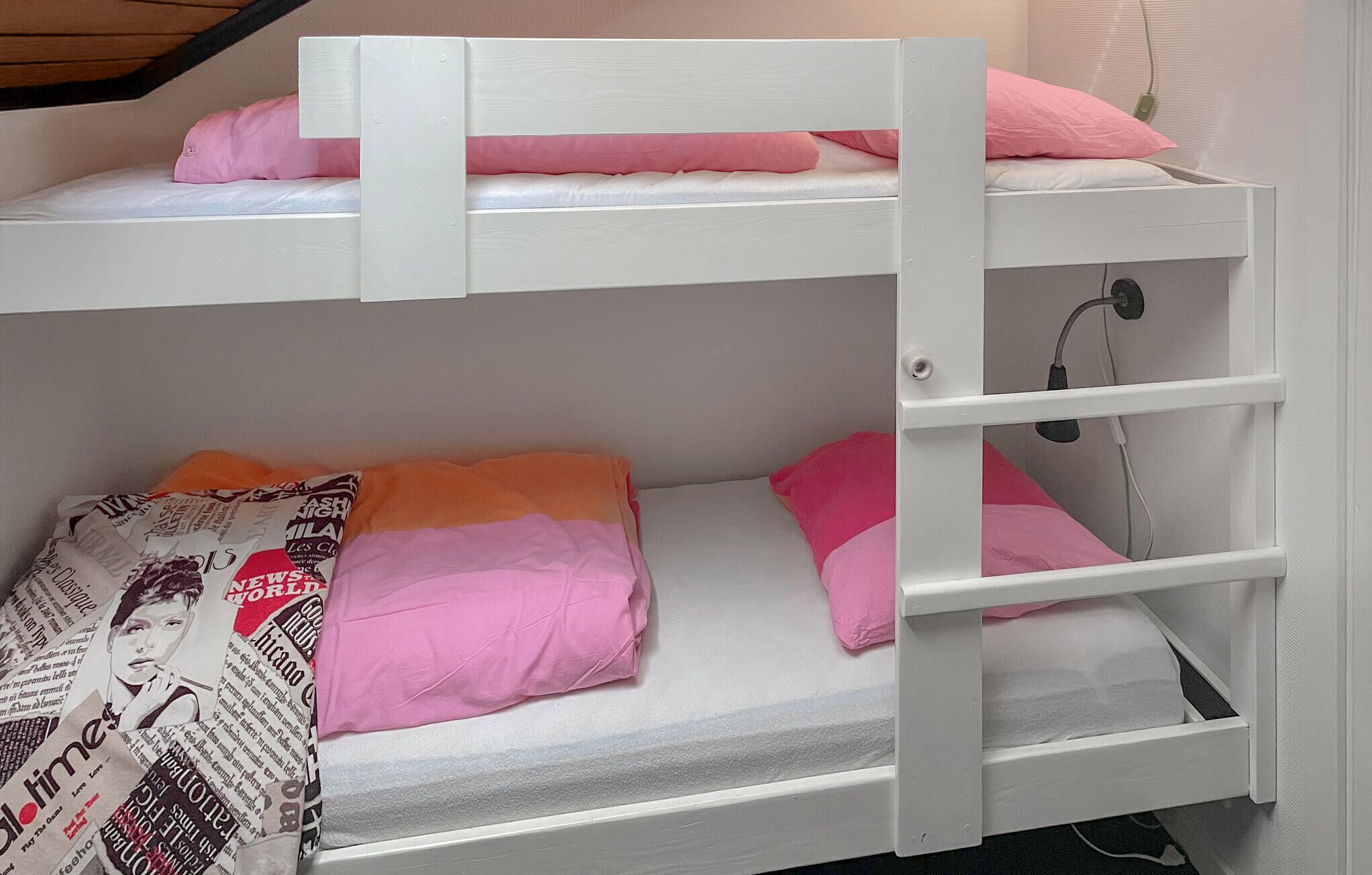 Ein Stockbett mit pinkener und orangener Bettwäsche, einem weißen Kopfteil und einem kleinen Regal.