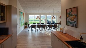 A modern kitchen with wooden cabinets, a dining area with a large table and chairs, and a view of the outdoors through large windows. - Amazing home in Hadsund with sauna (Hadsund)