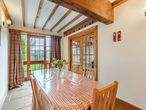 Un coin repas avec une table dressée pour six personnes, des chaises en bois, un vase rempli de fleurs et une vue sur l’extérieur grâce à une grande fenêtre agrémentée de rideaux.