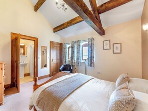 Un dormitorio con techo de madera, una cama con almohada estampada, una silla, una ventana con cortinas y una puerta que da a un baño.