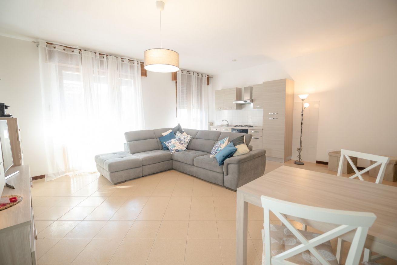 Een moderne woonkamer met een grijze hoekbank, een eettafel met witte stoelen en een keuken in de achtergrond.