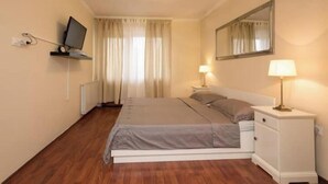Un dormitorio con cama, mesita de noche con lámpara, televisor montado en la pared y ventana con cortinas.