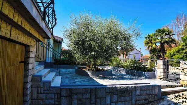 Un patio con una escalera de piedra que lleva a una puerta de madera, rodeado de follaje y un cielo azul despejado.