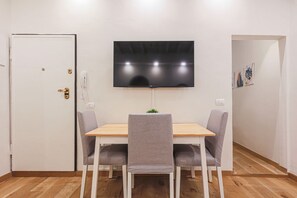 A dining area with a wooden table, two grey chairs, a flat-screen TV mounted on the wall, and a door with a brass handle.