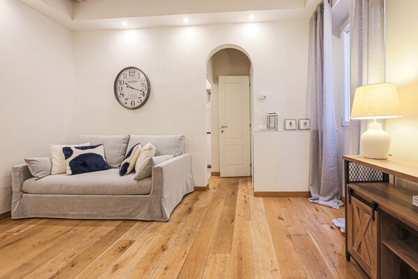 A living room with a sofa, a clock, a wooden floor, a door, and a lamp.