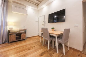 A room with a wooden floor, a TV mounted on the wall, a desk with chairs, and an air conditioning unit.