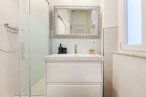 A bathroom with a glass shower enclosure, a white sink with a mirror above it, and a window to the right.