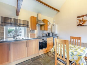 A modern kitchen with wooden cabinets, a built-in oven, a dishwasher, and a window with a view of greenery. - Eight Byre - UK48913 (Allonby)