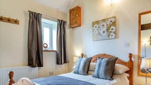 Ein Schlafzimmer mit hölzernem Bettgestell, einem Fenster mit Vorhängen, einem Nachttisch mit Lampe und einem an der Wand befestigten Spiegel.