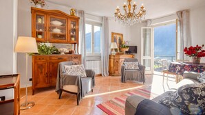A spacious living room with a large window, a chandelier, and a balcony with a view.