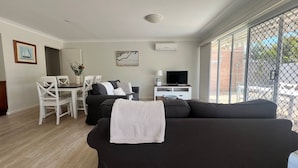 Un soggiorno con un divano nero, un tavolo da pranzo con sedie bianche, una TV su un mobile e un'ampia finestra con vista all'esterno.