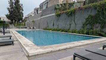 Ein rechteckiger Swimmingpool mit Fliesenrand, umgeben von Liegestühlen und einer Betonmauer mit Bepflanzung.