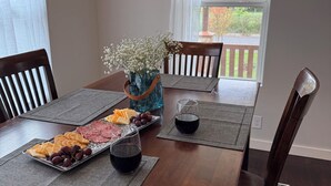Una mesa de comedor dispuesta con una variedad de quesos, galletas y uvas, acompañada de dos copas de vino tinto, en una habitación con muebles de madera y vistas al exterior.