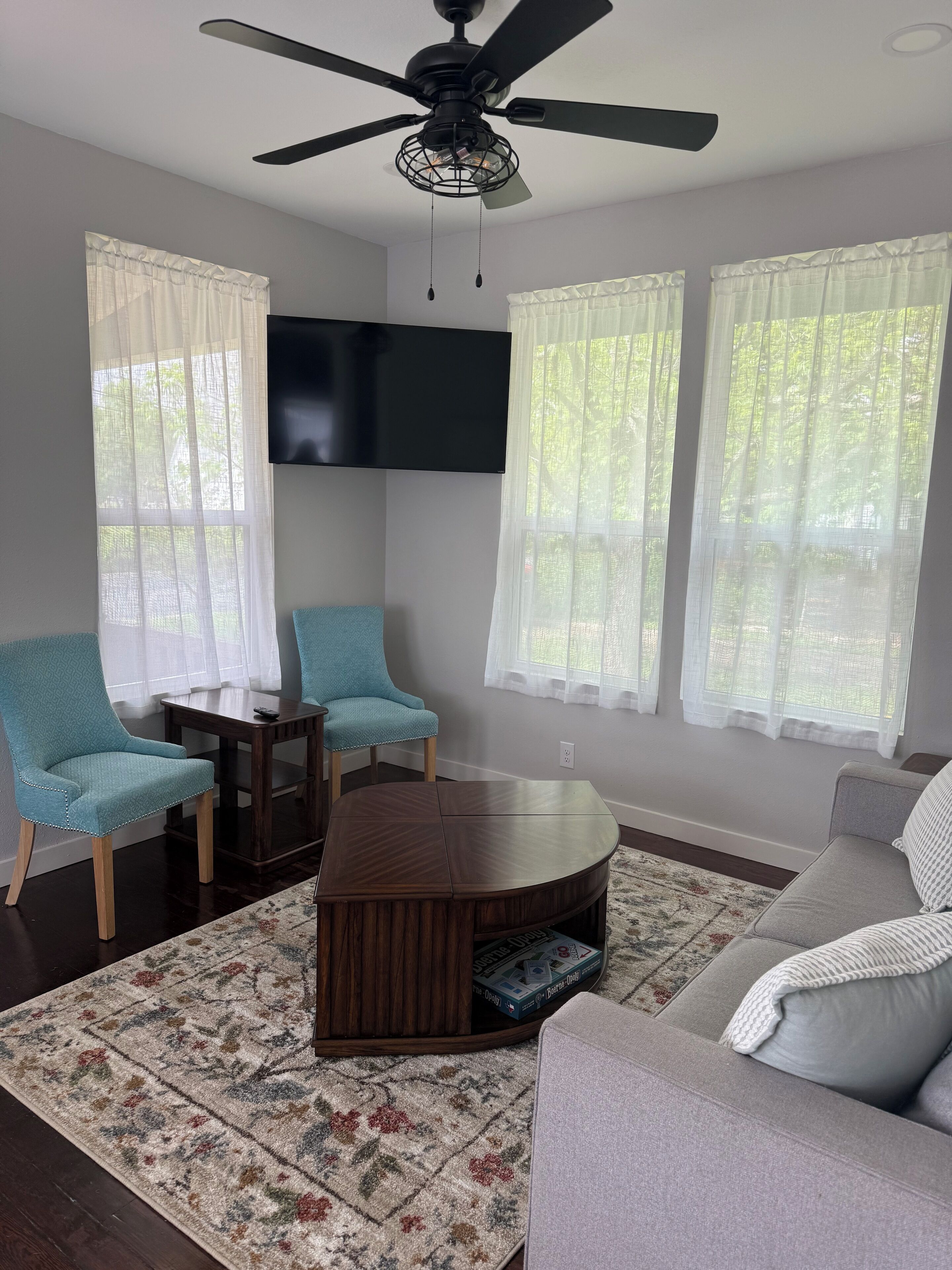 Ein Wohnzimmer mit Deckenventilator, zwei hellblaue Sessel, einem braunen Couchtisch, einem grauen Sofa und einem flachbildfernseher an der Wand.