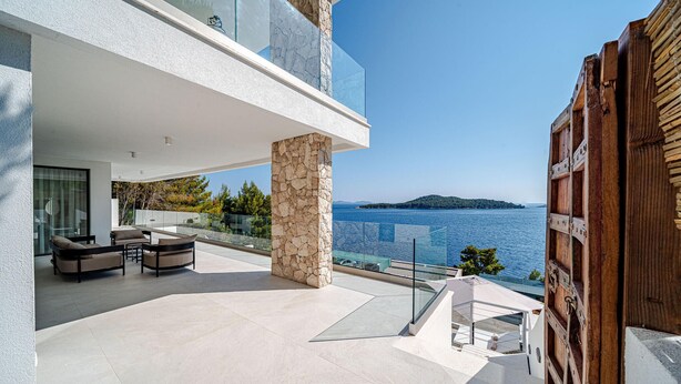 Un espace de vie extérieur moderne, agrémenté d’un pilier en pierre, d’une balustrade en verre et offrant une vue sur la mer.