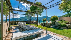 Un patio al aire libre con una pérgola, sillones de descanso y vistas a montañas y un lago.
