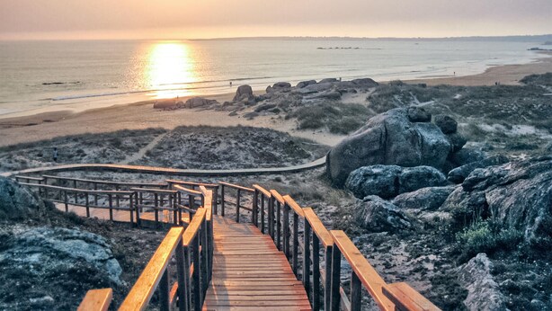 A wooden boardwalk leading to a beach with large rocks and a sunset in the background.