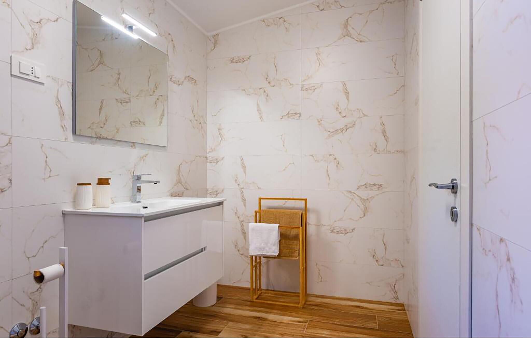 Une salle de bain avec un mur en marbre, un plancher en bois, un meuble-lavabo blanc avec un miroir et un porte-serviettes.