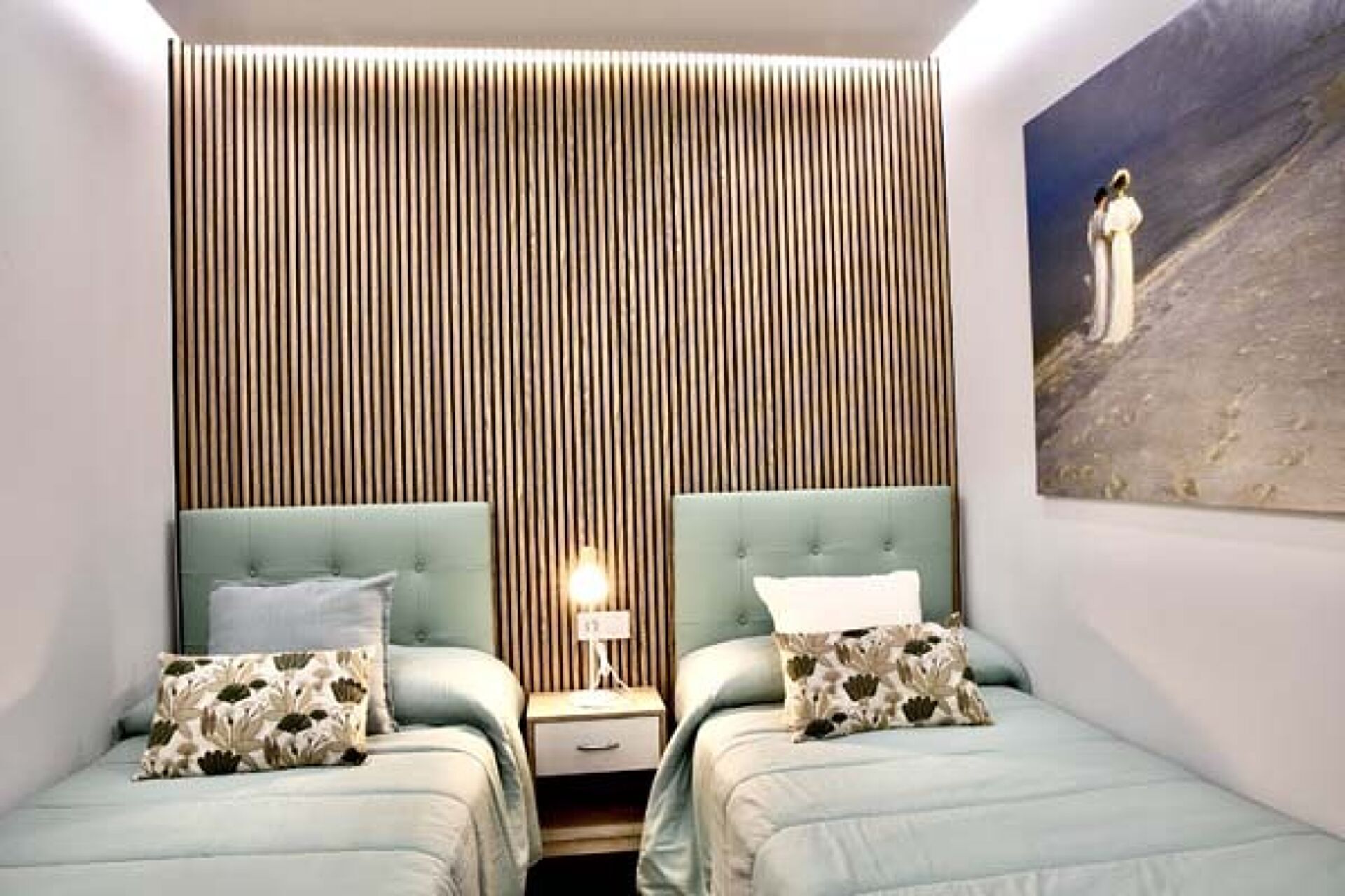 Un dormitorio con dos camas, una pared de listones de madera, una mesita de noche con lámpara y un cuadro en la pared.