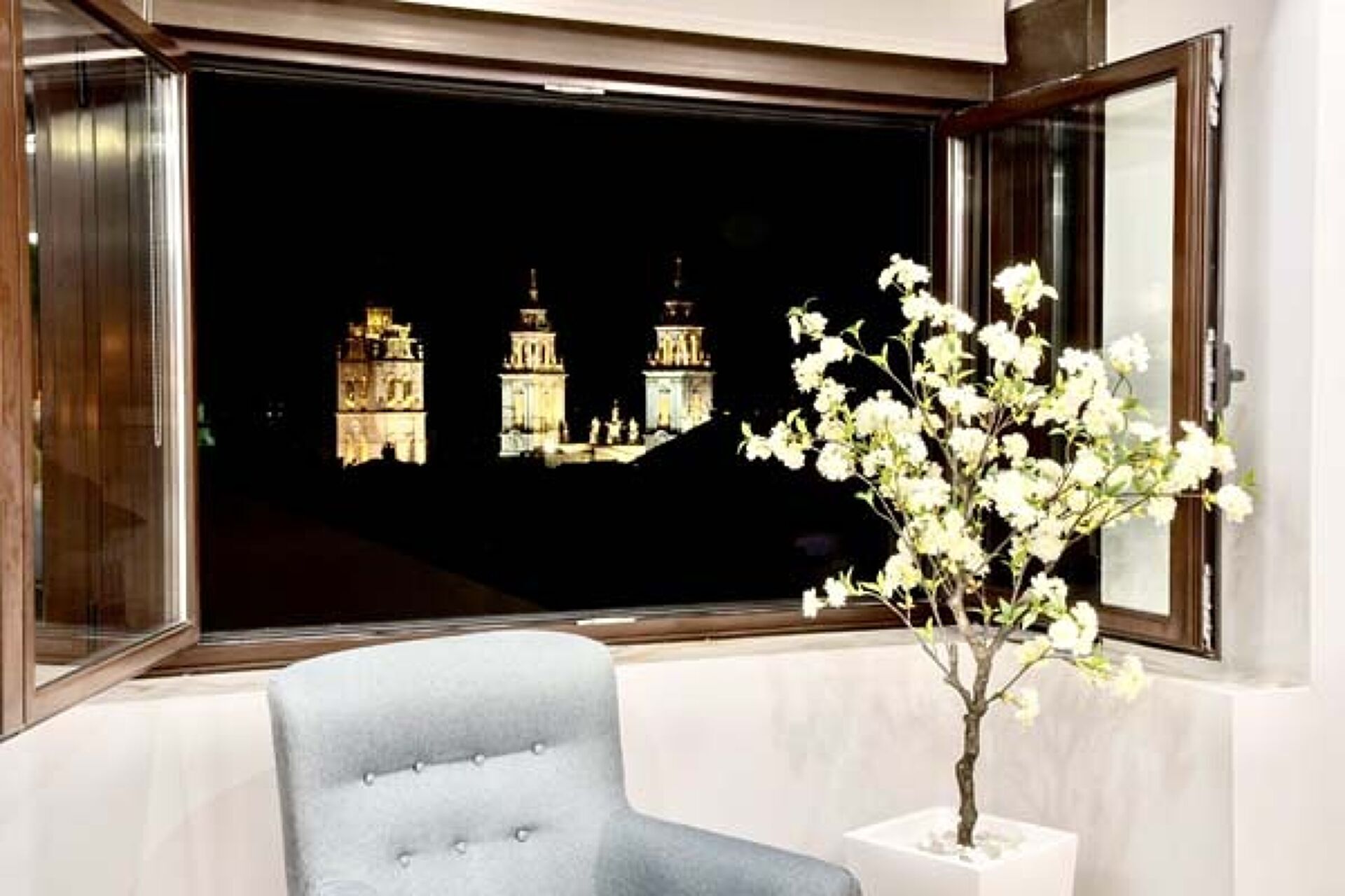A room with a window overlooking a cityscape at night, a white chair, and a potted plant with white flowers.