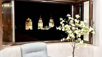A room with a window overlooking a cityscape at night, a white chair, and a potted plant with white flowers.