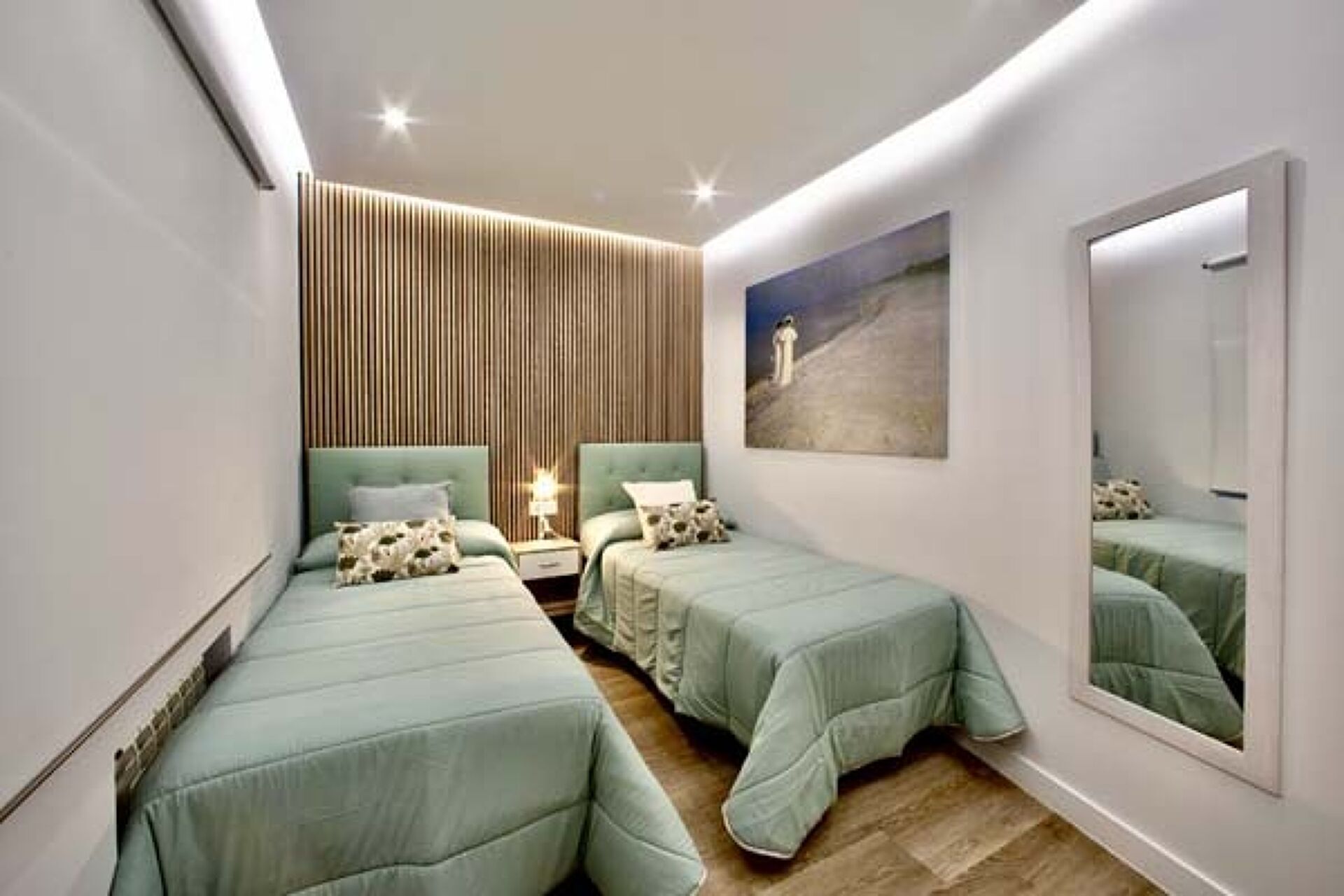 Un dormitorio con dos camas individuales, una mesita de noche con lámpara, un cuadro en la pared y un espejo grande.