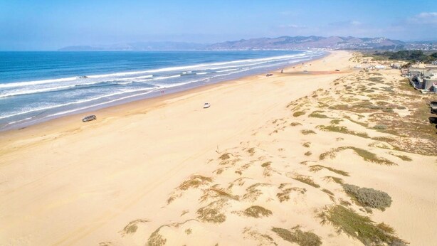 Una extensa playa arenosa con olas suaves, algunos vehículos y colinas a lo lejos.