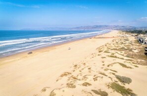 Una extensa playa arenosa con olas suaves, algunos vehículos y colinas a lo lejos.