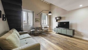 Uma sala de estar com sofá, uma mesa de jantar com cadeiras, uma TV em um móvel e uma escada.