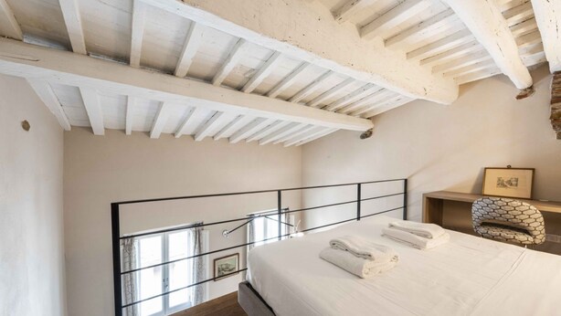 Quarto com teto de madeira, cama com roupa de cama branca, corrimão de metal e uma pequena mesa com moldura de quadro.
