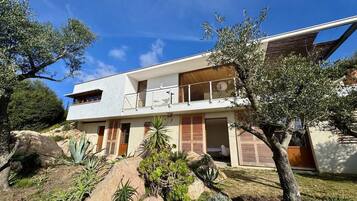 Une maison moderne à deux étages, dotée d’un balcon et entourée d’un aménagement paysager naturel comprenant des rochers et des arbres.