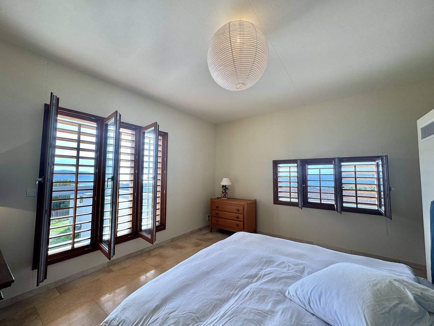 A bedroom with a bed, a nightstand, a lamp, and a view of the sea through a window.