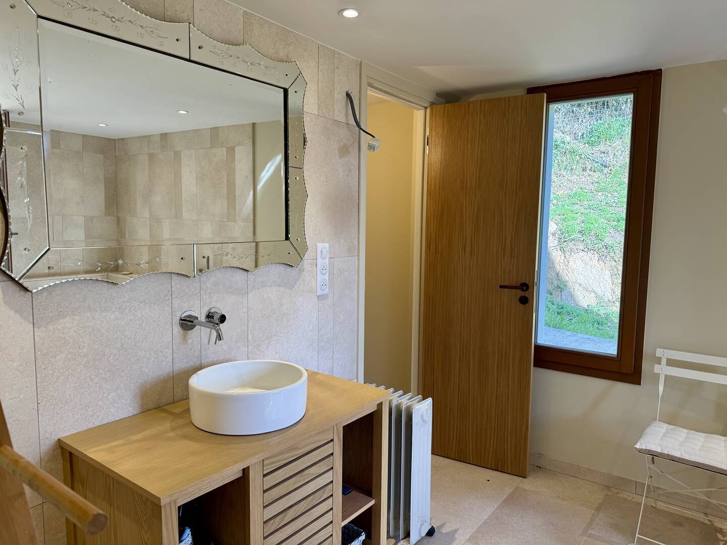 A bathroom with a large mirror, a round sink, a wooden vanity, a radiator, and a door leading to a garden.