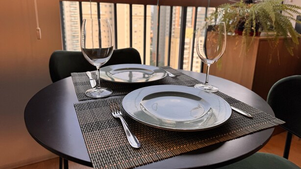 A dining table set with plates, cutlery, and wine glasses, positioned on a balcony with a view of high-rise buildings. - Studio Brooklin 1min from the subway and close to the consulate (Brooklin São Paulo)