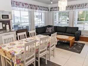 A modern living room with a dark grey sofa, white chairs, a dining table with floral tablecloths, and large windows with curtains.
