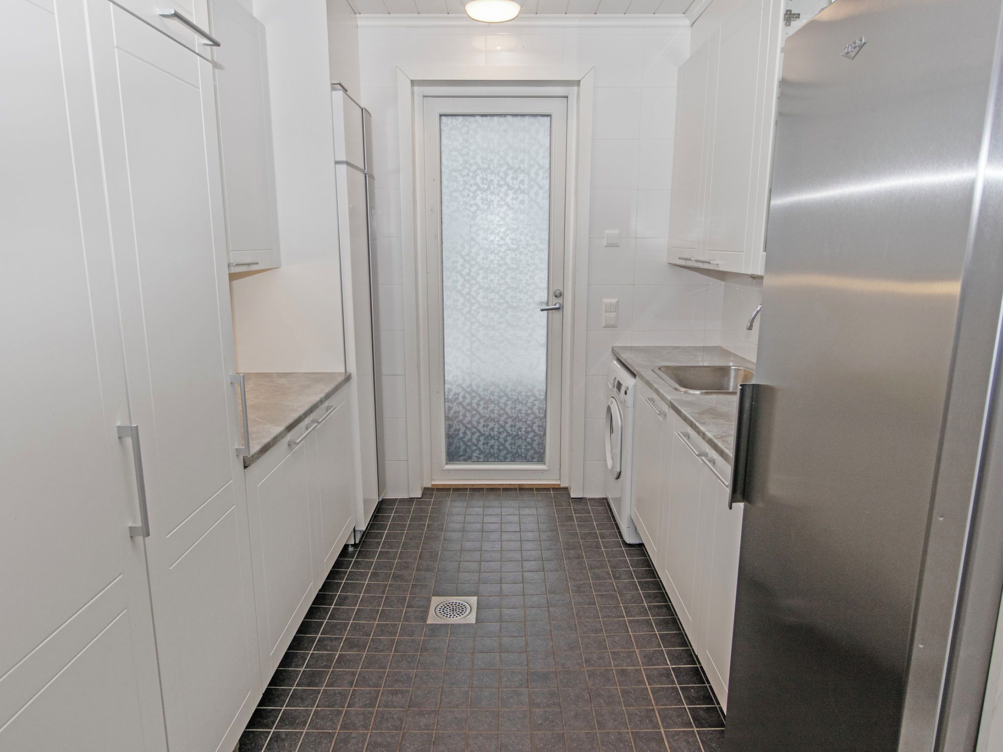 A narrow kitchen with white cabinets, a stainless steel refrigerator, and a tiled floor.